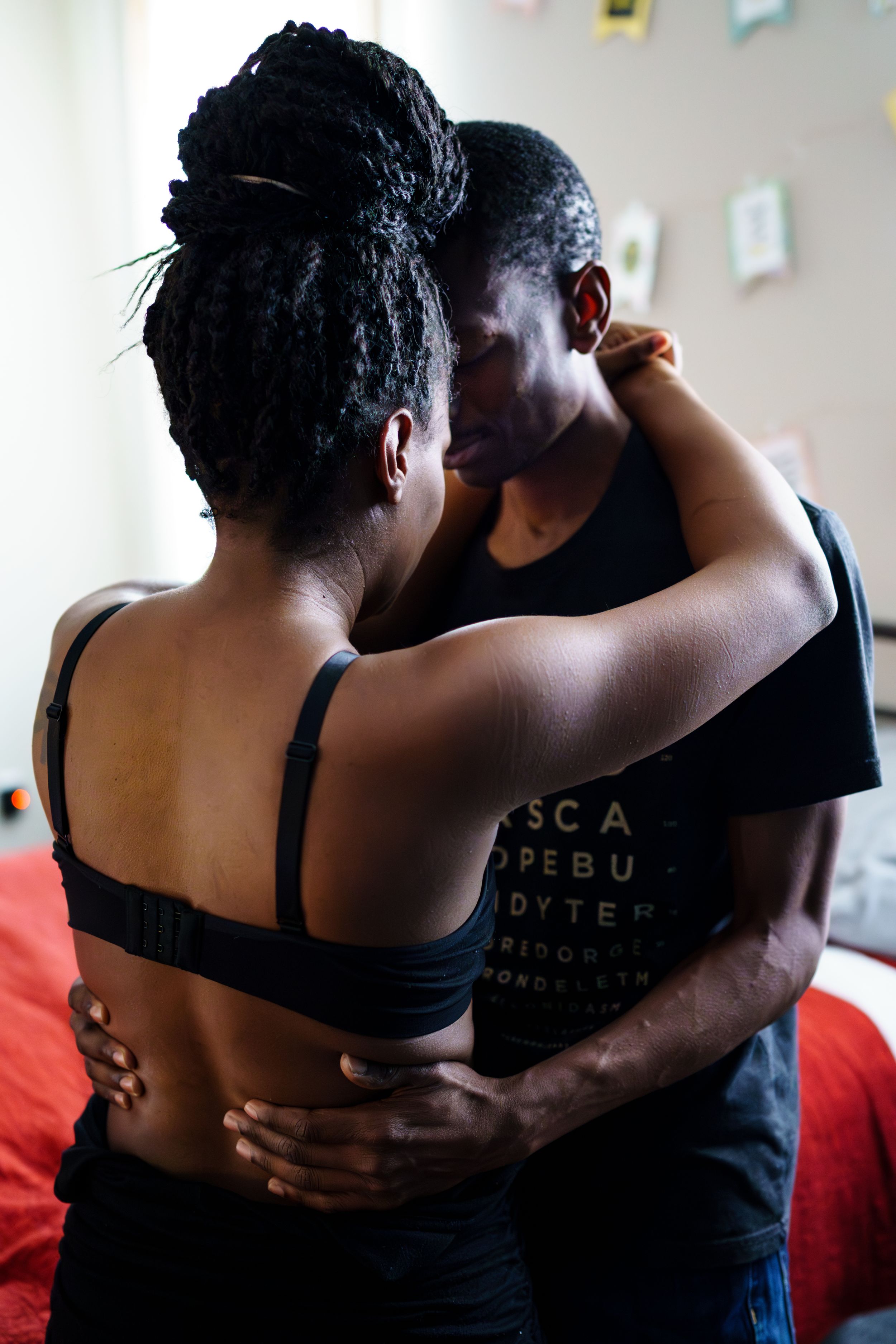 A couple shares an intimate embrace while standing beside a red bed.