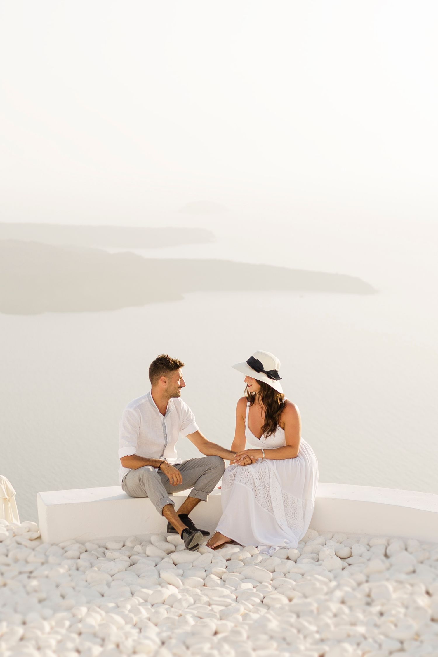 A couple in white clothing sits on a ledge overlooking a misty coastal landscape.