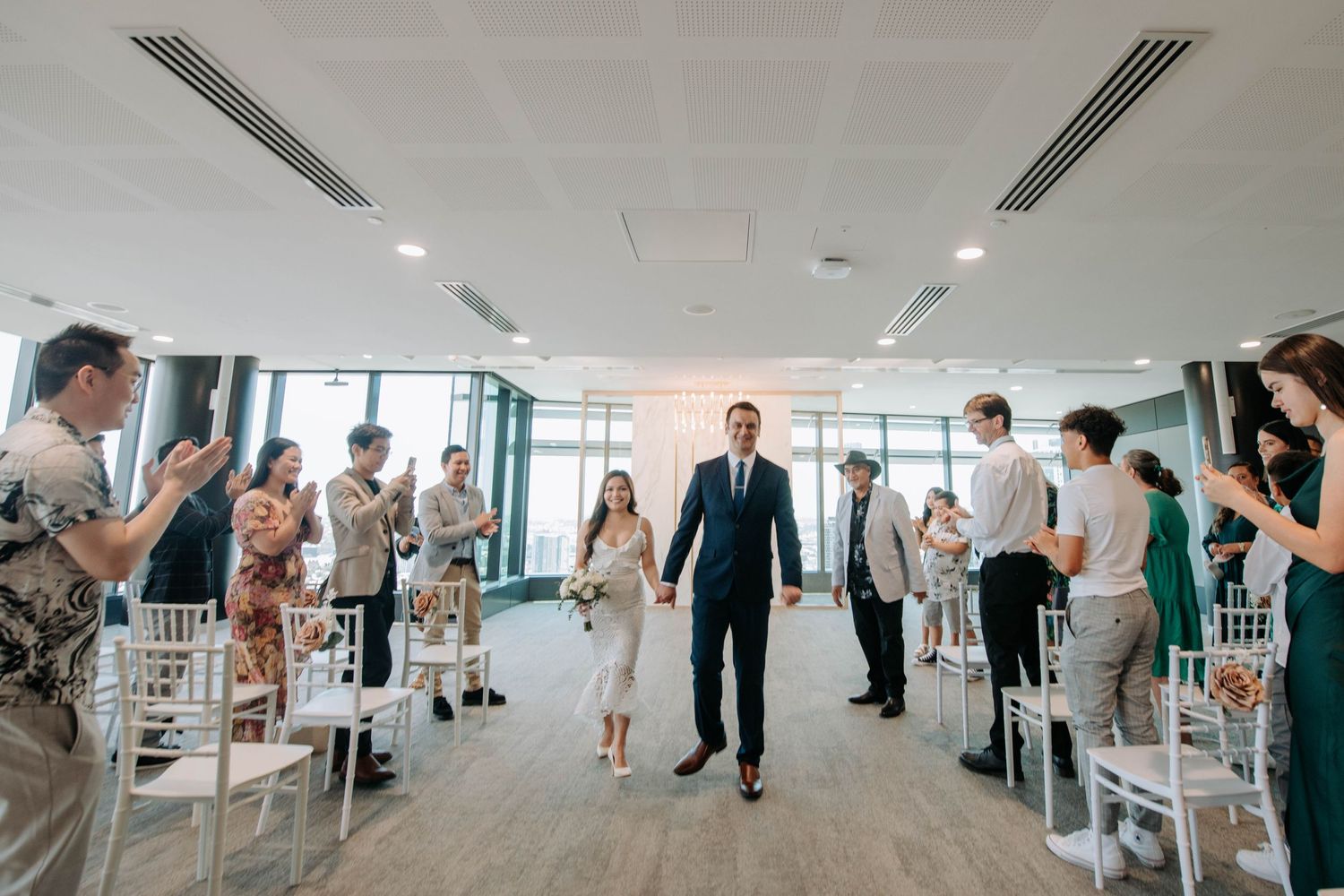 Newlyweds walking down the aisle while guests celebrate in modern bright venue.
