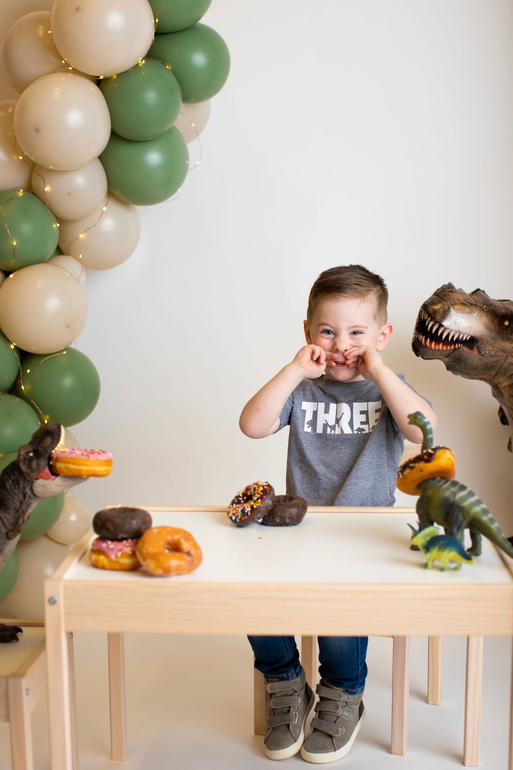 Dinosaur themed birthday setup with green and beige balloons and donuts on white table.