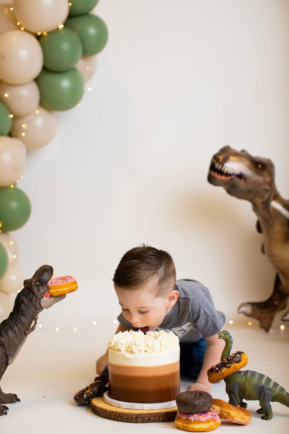 Child leaning over birthday cake with dinosaur decorations and balloon arch.