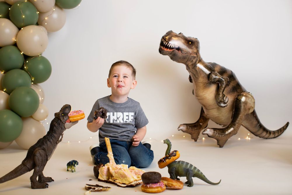 Fun dinosaur themed photo setup with toys, donuts and green balloons against white background.