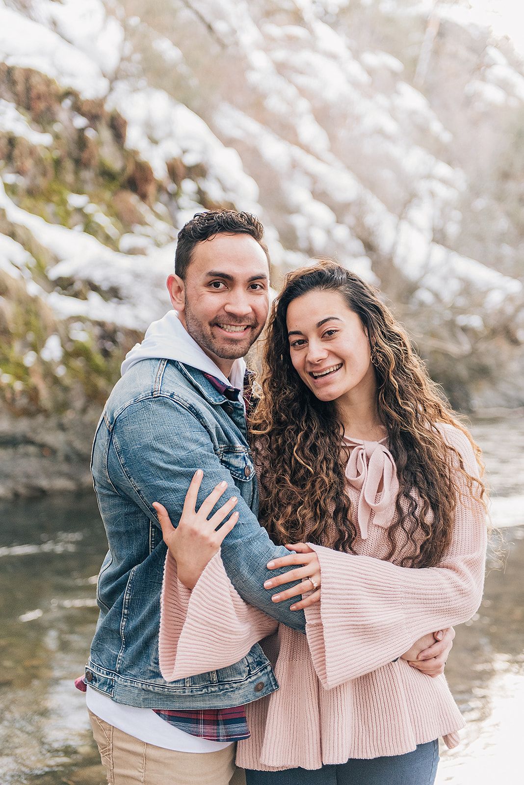 A couple embraces in winter by a stream, wearing a denim jacket and pink sweater against snowy backdrop.