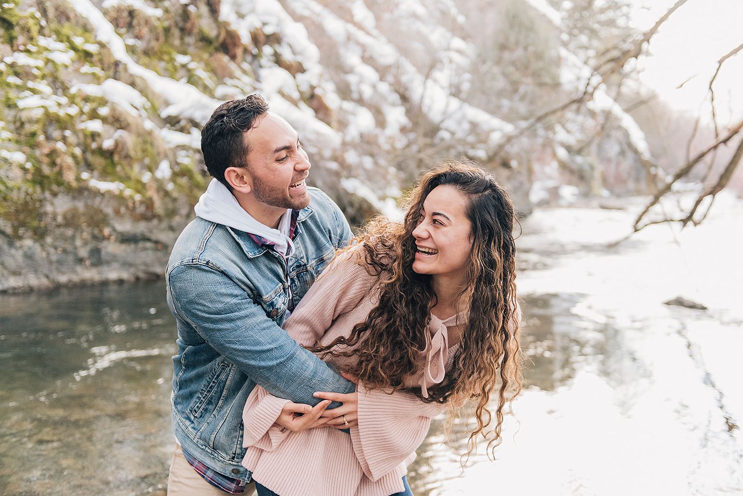 A couple shares a playful moment near a winter stream wearing casual denim and pink.