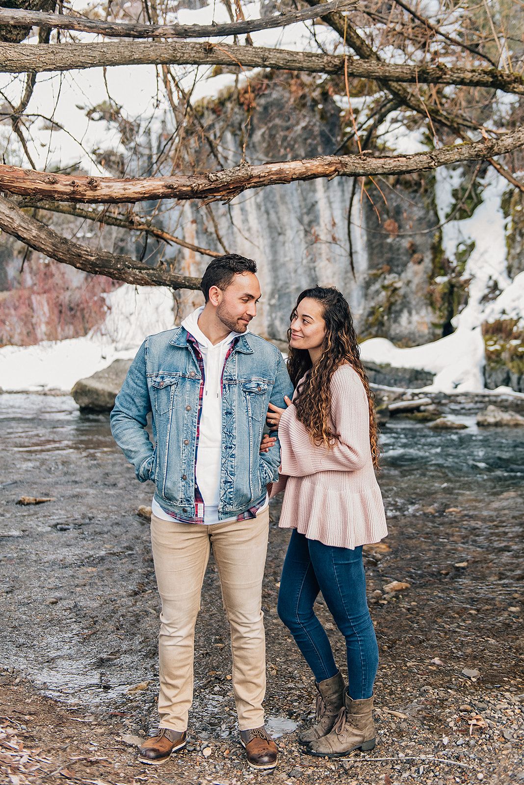 A couple laughs together by a flowing stream in winter wearing casual outfits.
