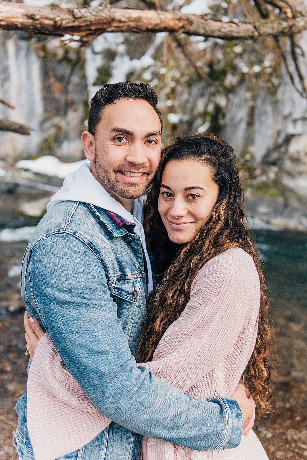 A couple embraces while wearing matching pink and denim outfits near a waterfall.