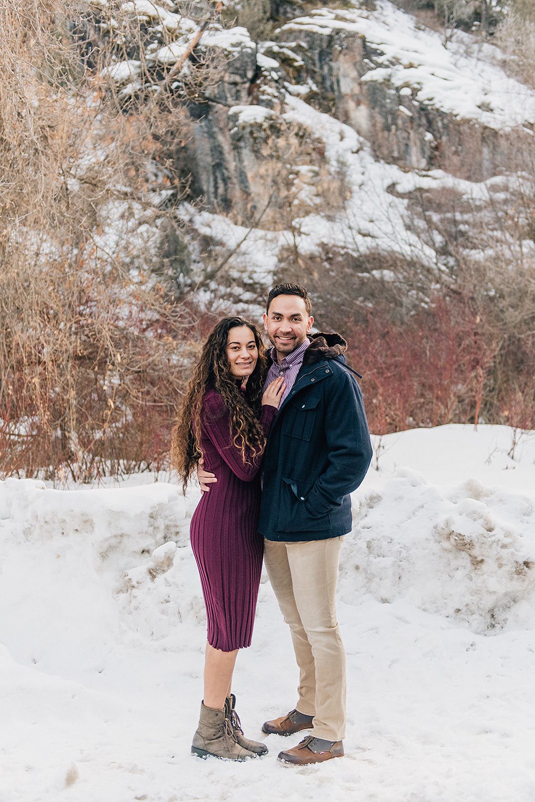 A winter engagement photo of a couple standing in front of a snow-dusted mountain cliff.