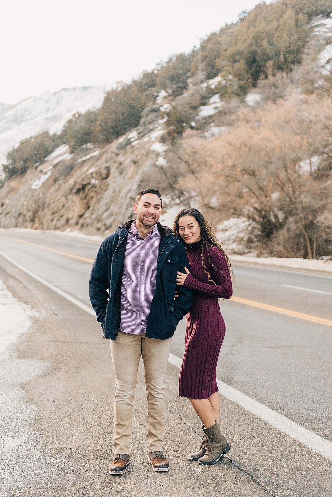 A couple poses together on a mountain road with snowy cliffs in the background.