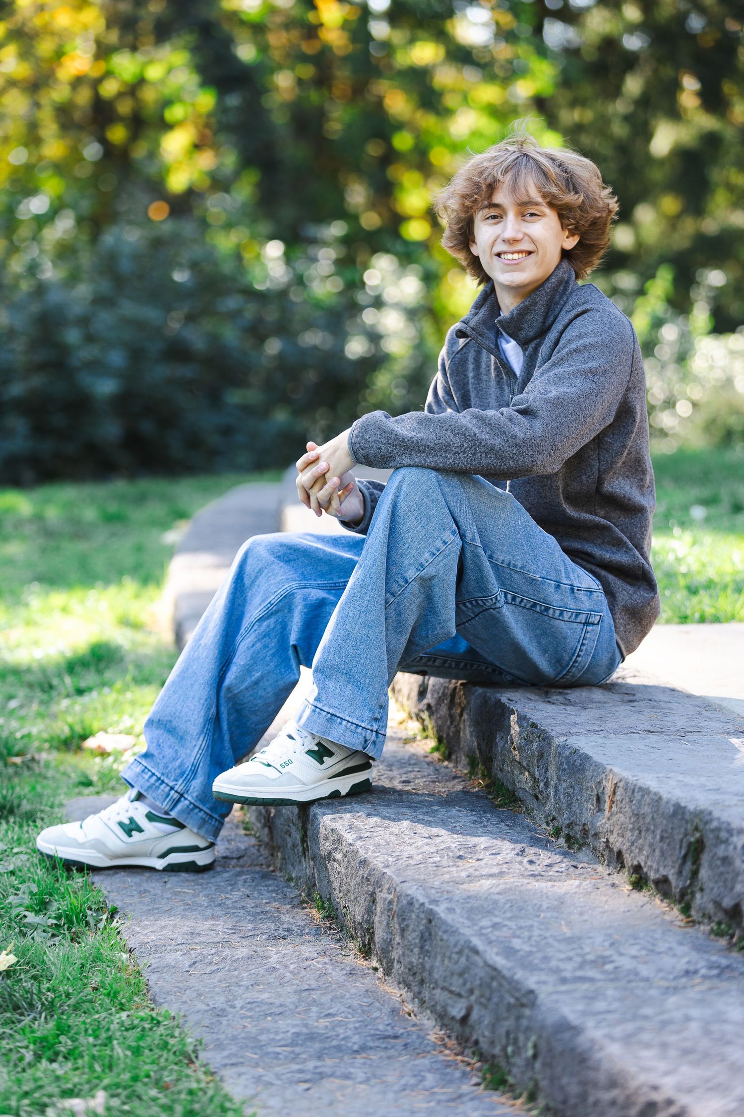 A person in jeans and gray sweater sits casually on stone steps in a sunny park setting.