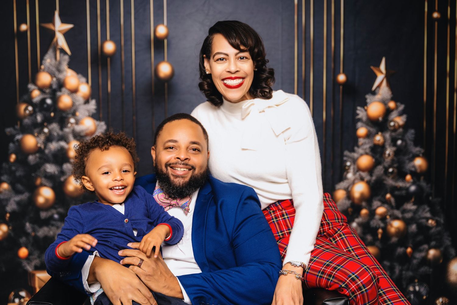 A family poses for a holiday portrait with decorated Christmas trees and gold lights behind them.