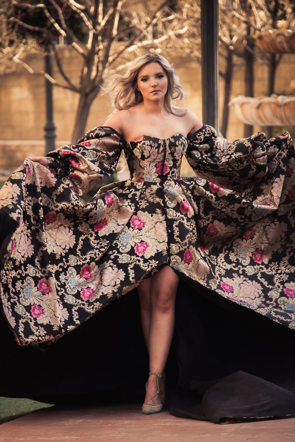 Blonde woman in a dramatic floral brocade ball gown with wide sleeves posing outdoors.