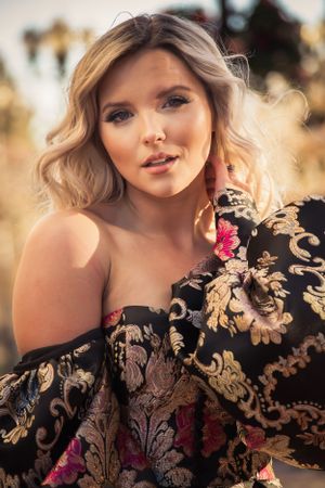 Blonde woman in off-shoulder black floral brocade gown poses outdoors in warm golden light.