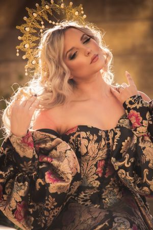 Blonde woman wearing gold crown poses in black pink and gold floral brocade off-shoulder gown.