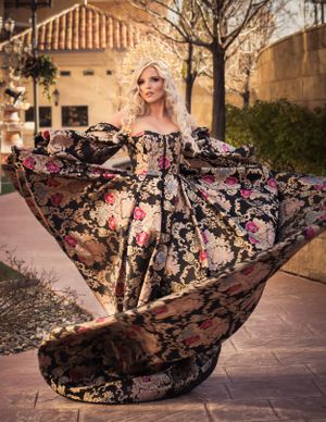 Blonde woman twirls in stunning black gold and pink floral brocade ballgown wearing ornate crown outdoors.