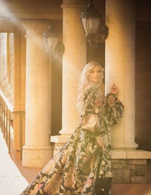 Elegant blonde woman in black floral brocade gown stands among classical columns in golden sunlight.