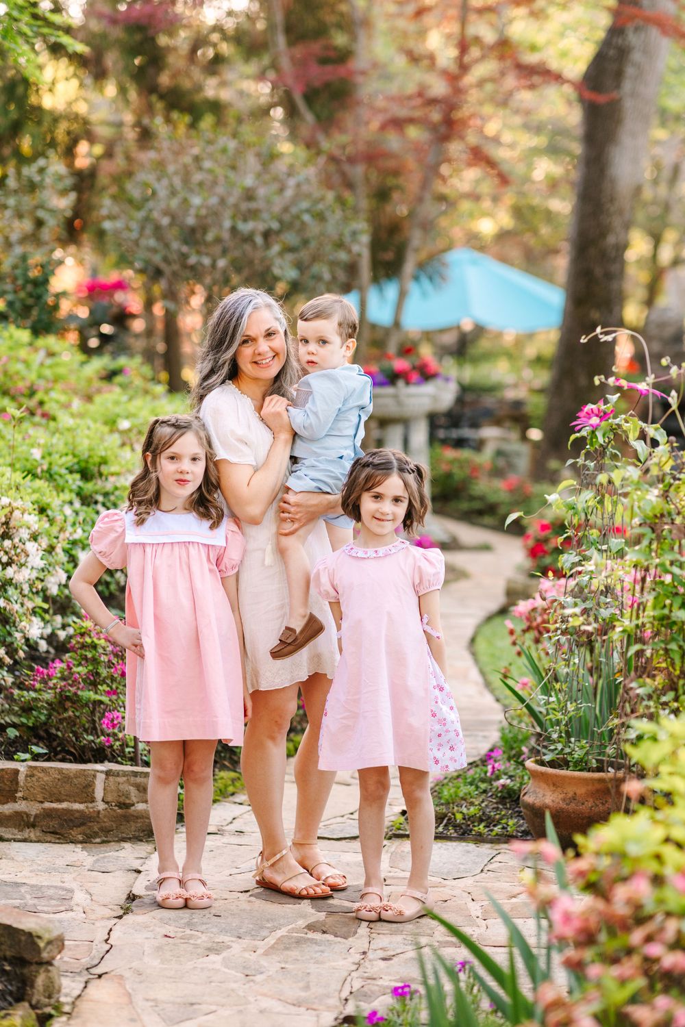 A family group stands on a stone garden path surrounded by blooming flowers and greenery.