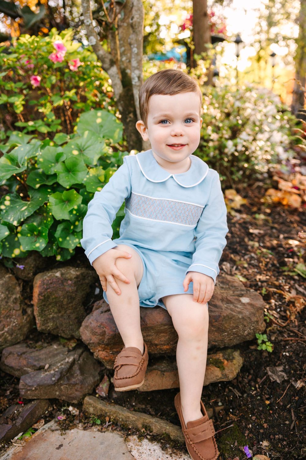 A young child in a light blue outfit sits on stone steps in a garden setting with blooming flowers at sunset.