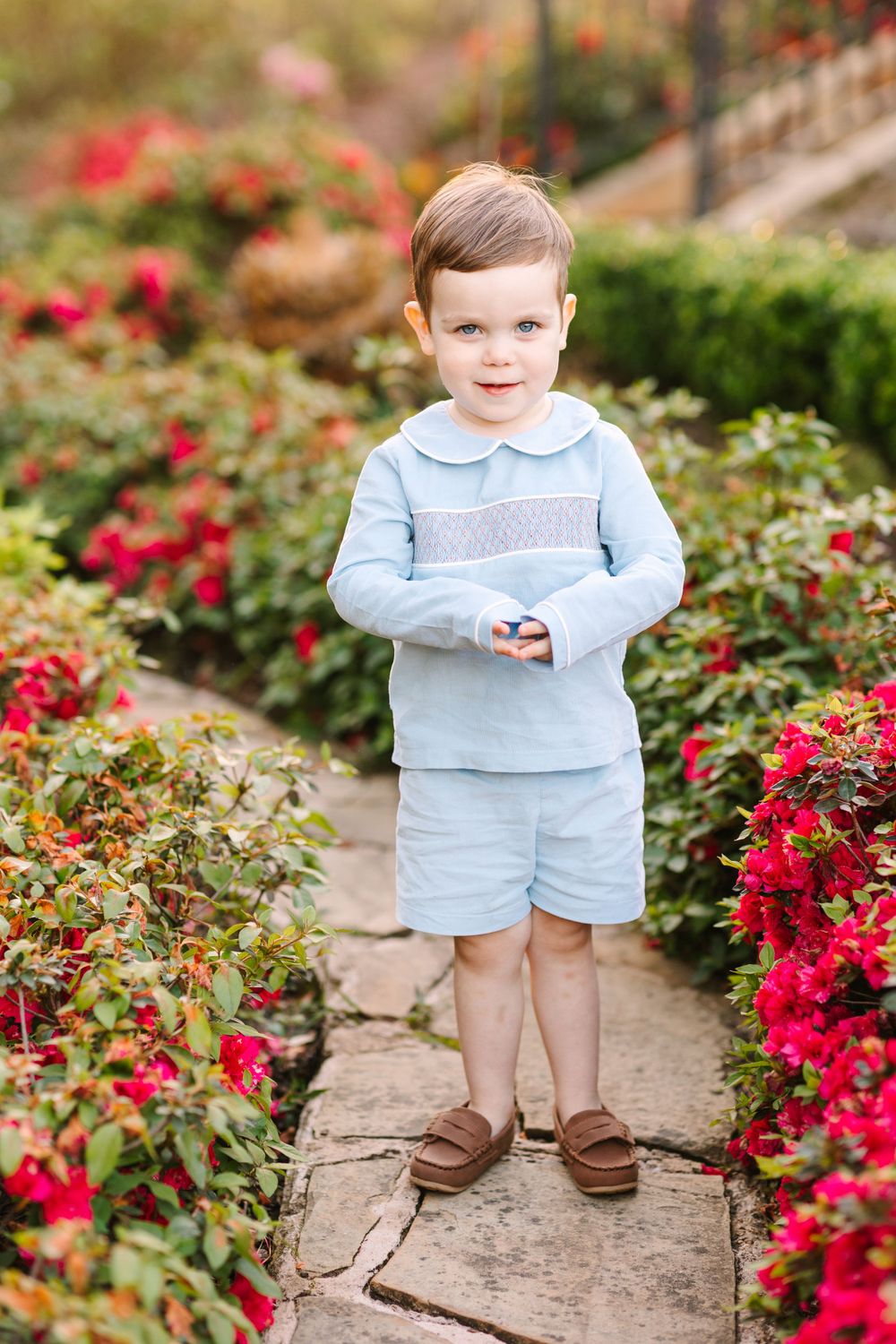 Young child in light blue outfit and brown shoes standing on garden path with red flowers.