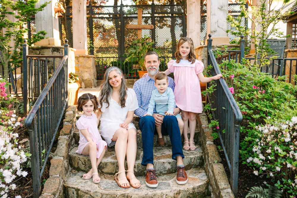 A family sits together on outdoor stone steps surrounded by lush greenery and blooming flowers.