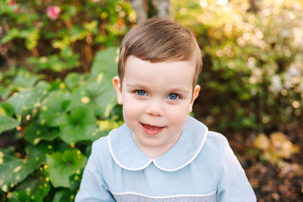 A cheerful child in a blue peter pan collar outfit poses in front of garden greenery.