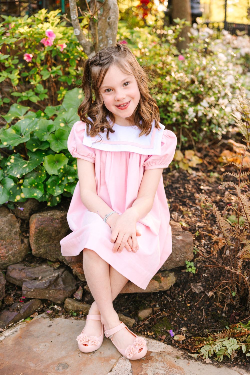 A child in a pink dress sits on stone steps surrounded by blooming flowers and greenery.