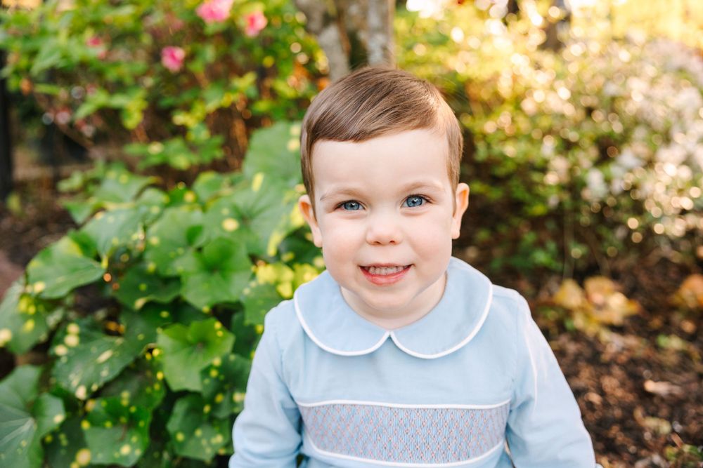 A close-up portrait of a young child in a blue collared outfit against a garden background.