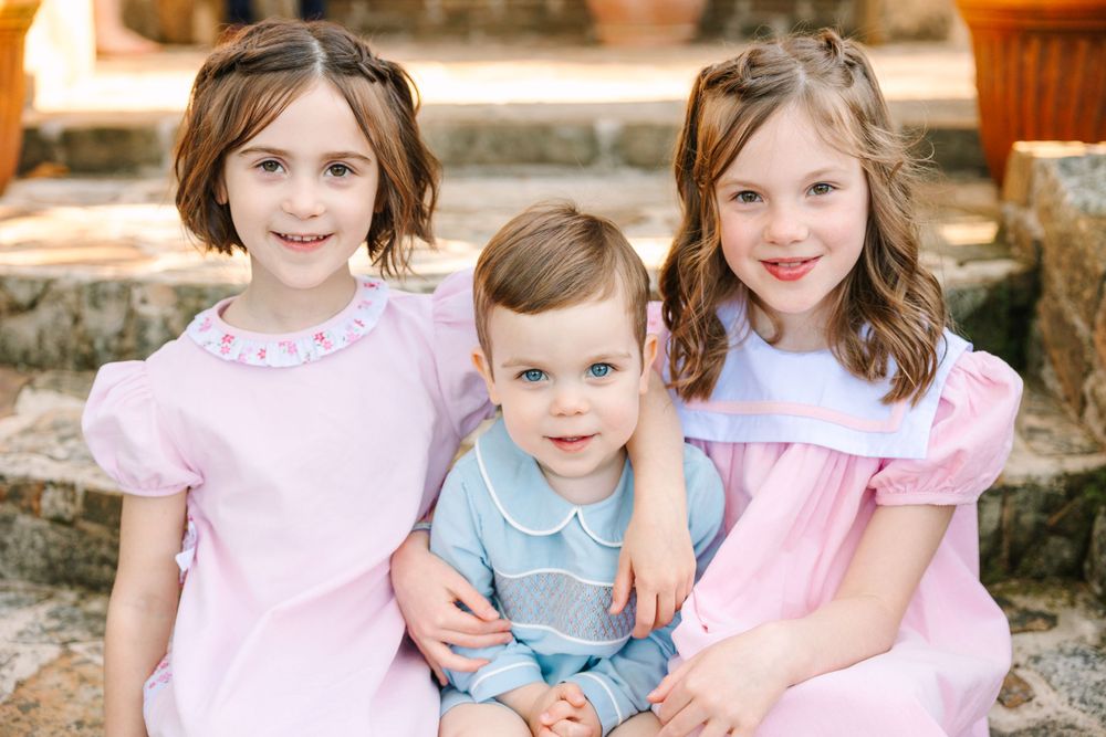 Three young children in pink and blue outfits sit together on stone steps in a garden setting.