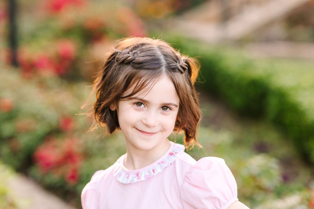 Child wearing pink dress poses in garden setting with red flowers.
