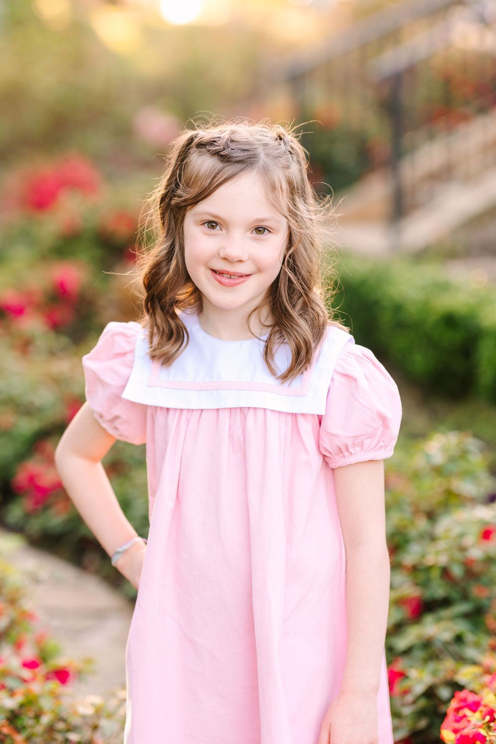 Child in pink puff sleeve dress standing in garden with blooming red flowers in background.