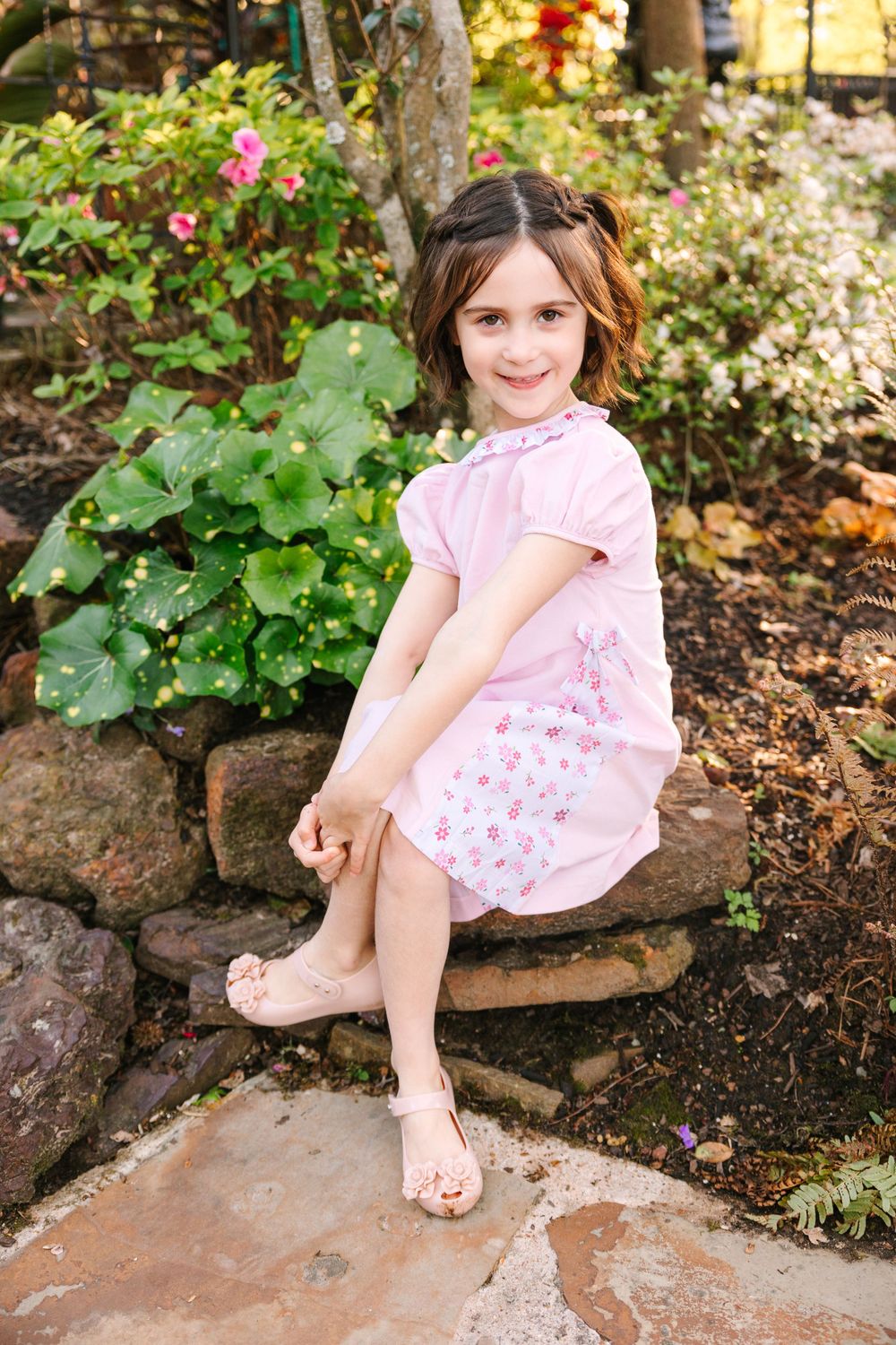 Child in pink dress sitting on garden stone wall surrounded by greenery.