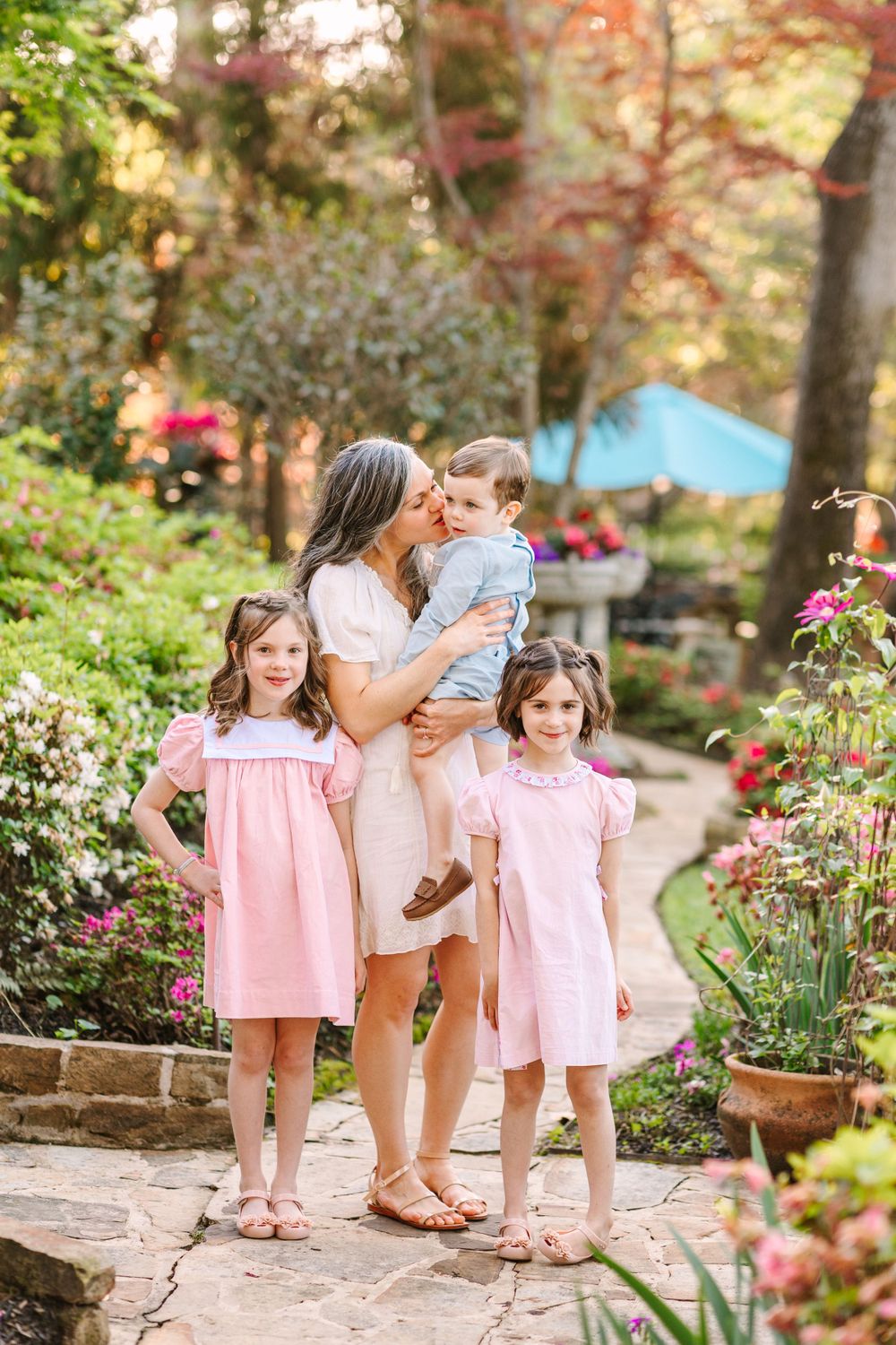 A garden portrait with three children in pink dresses and blue outfit against flowering landscaping.