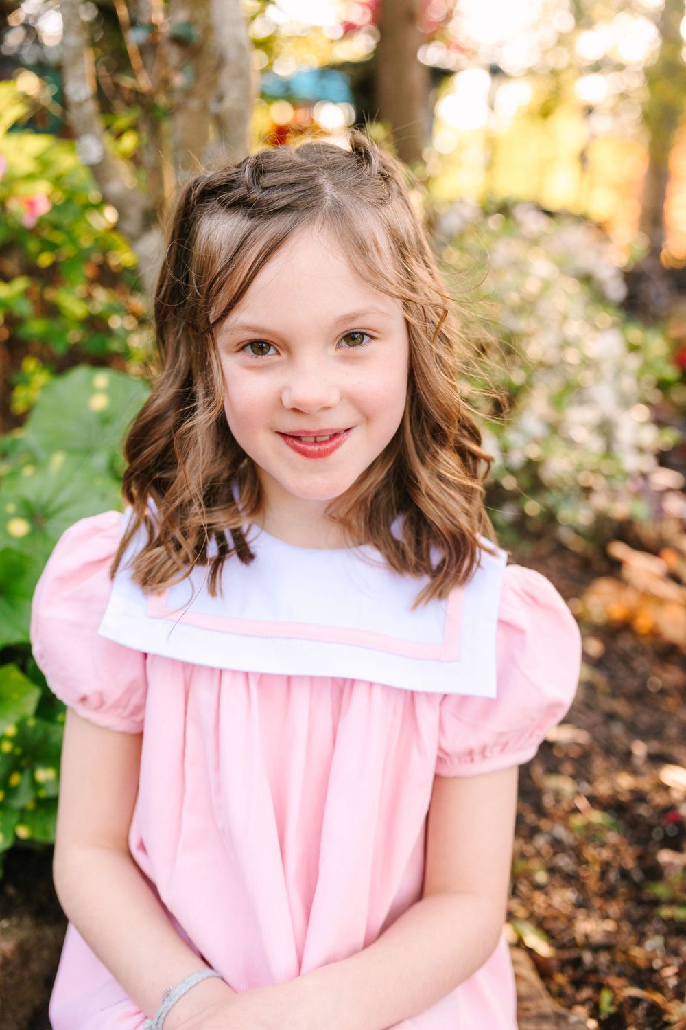 A child wearing a pink dress with puff sleeves poses against a colorful autumn garden background.