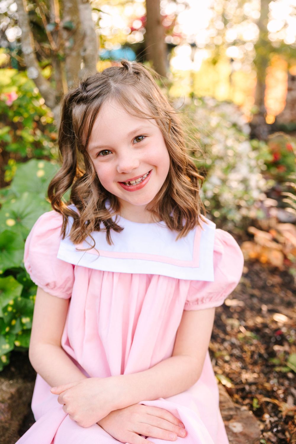 A child in a classic pink dress smiles while posing in a garden setting with flowering bushes.