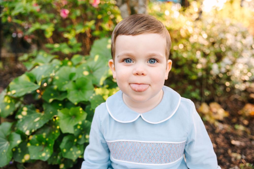 A smiling child in a blue and white collared outfit poses against green garden foliage.