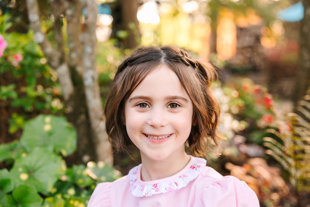 A child wearing a pink collared dress poses outdoors with blooming flowers and trees in the background.