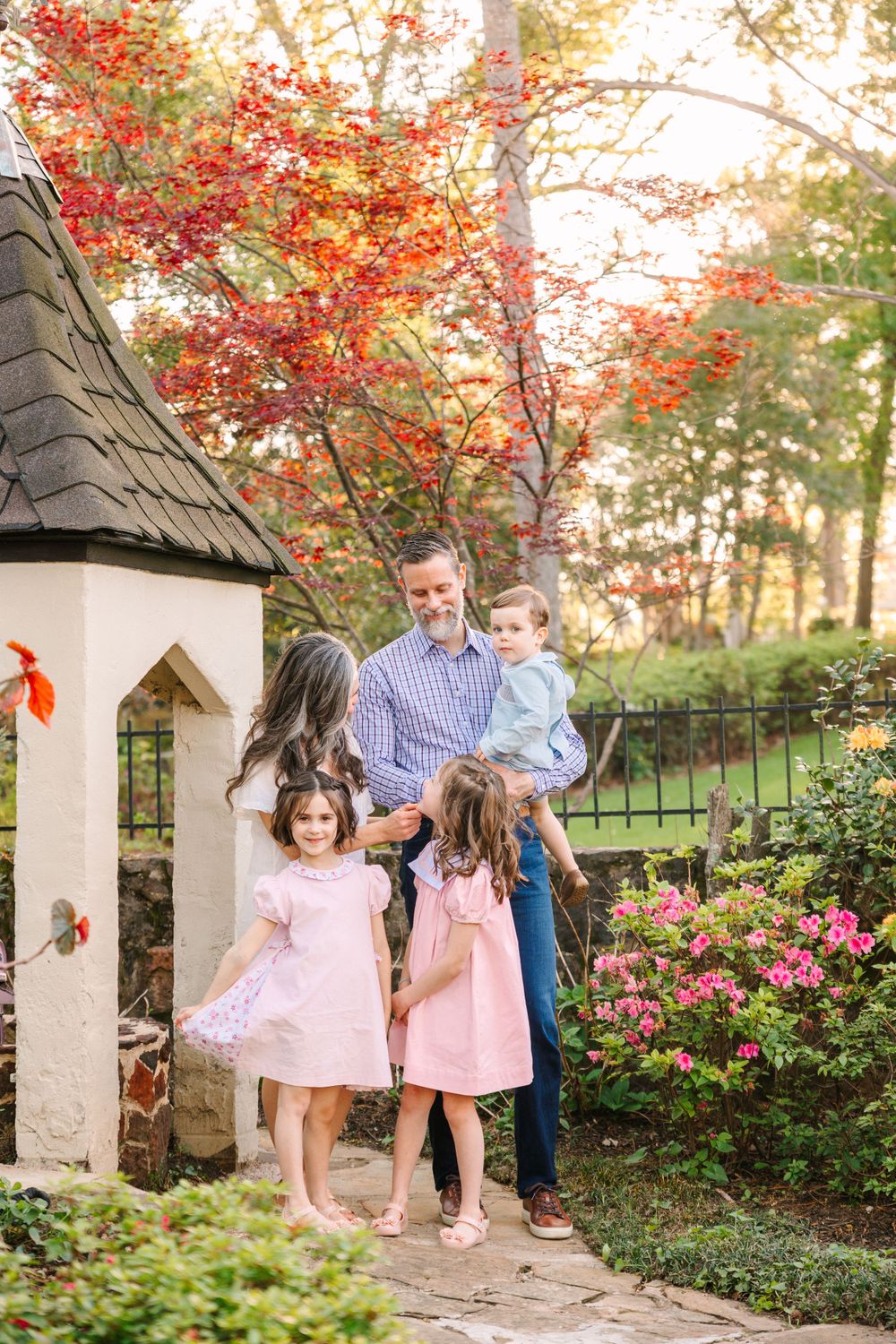 A family poses together near a small garden gazebo with vibrant red autumn foliage overhead.