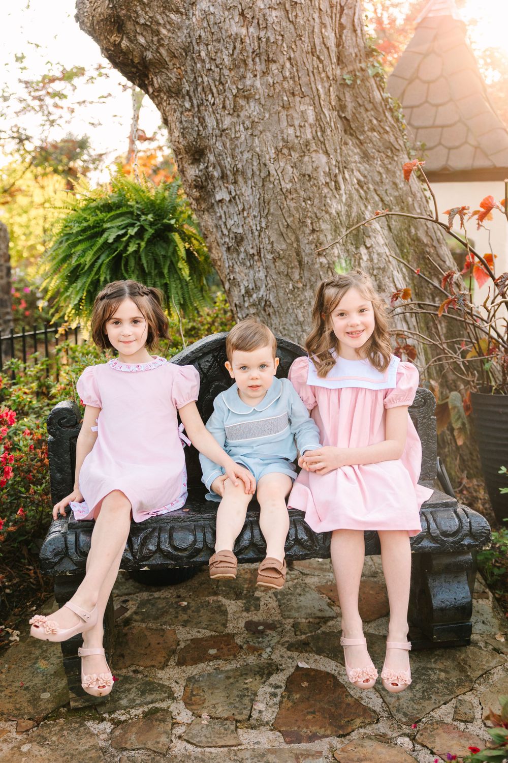Three children in coordinating outfits sit together on a stone bench under a large tree.