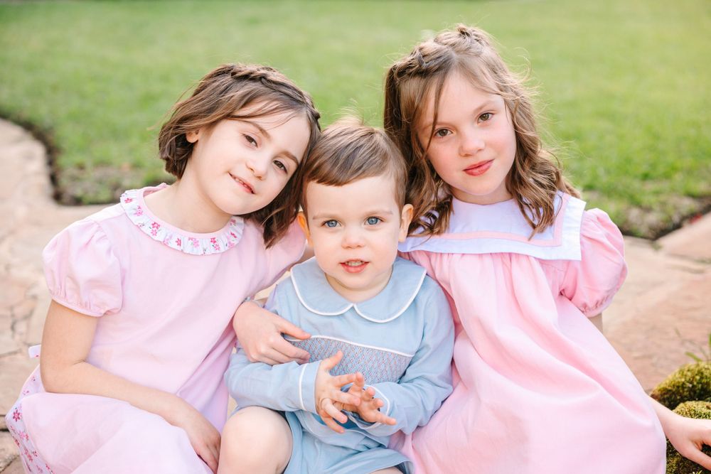 Three children in coordinating pink and blue outfits pose together outdoors on a sunny day.