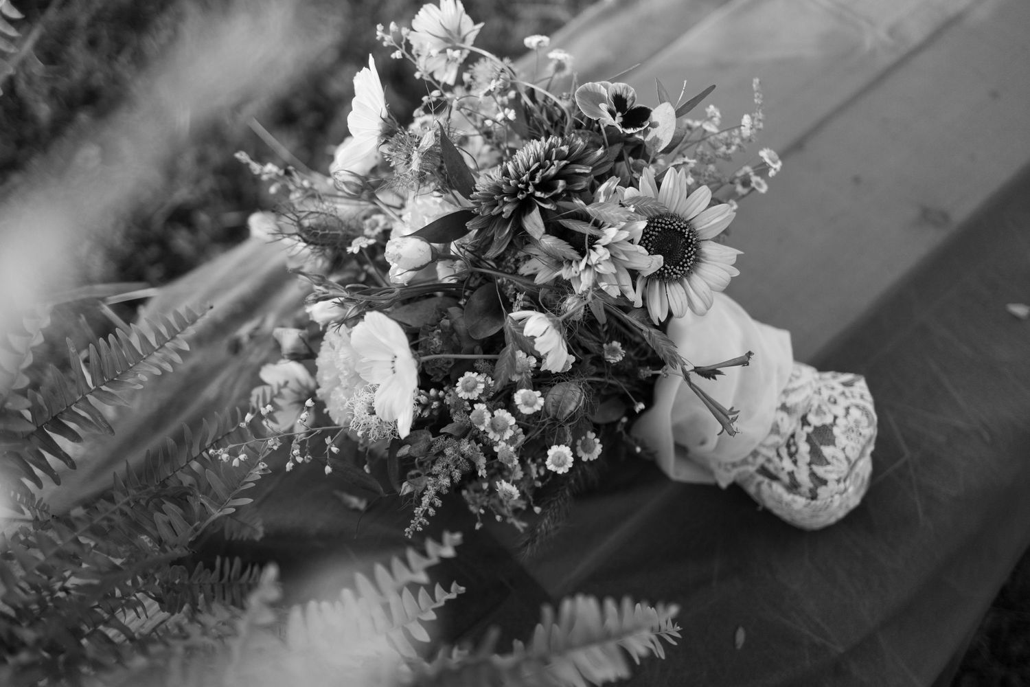Black and white photo of a floral bouquet with sunflowers and ferns on a wooden surface.