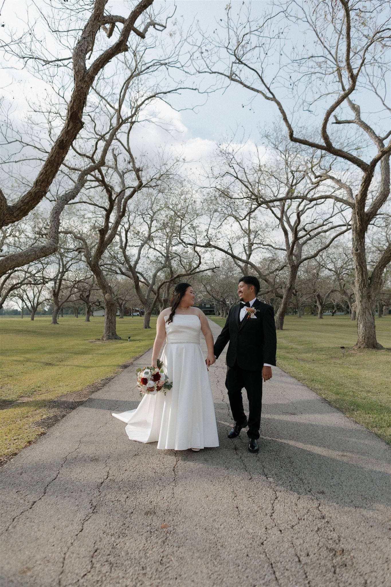 A couple walks hand in hand along a tree-lined pathway in an outdoor wedding setting.