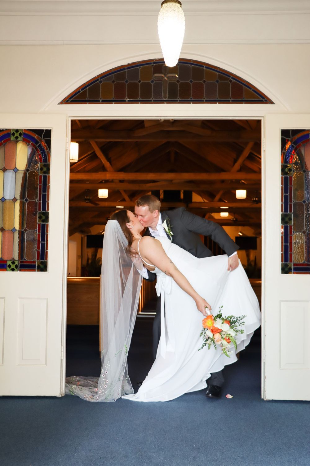 Wedding couple shares romantic dip pose in church hallway with stained glass windows.