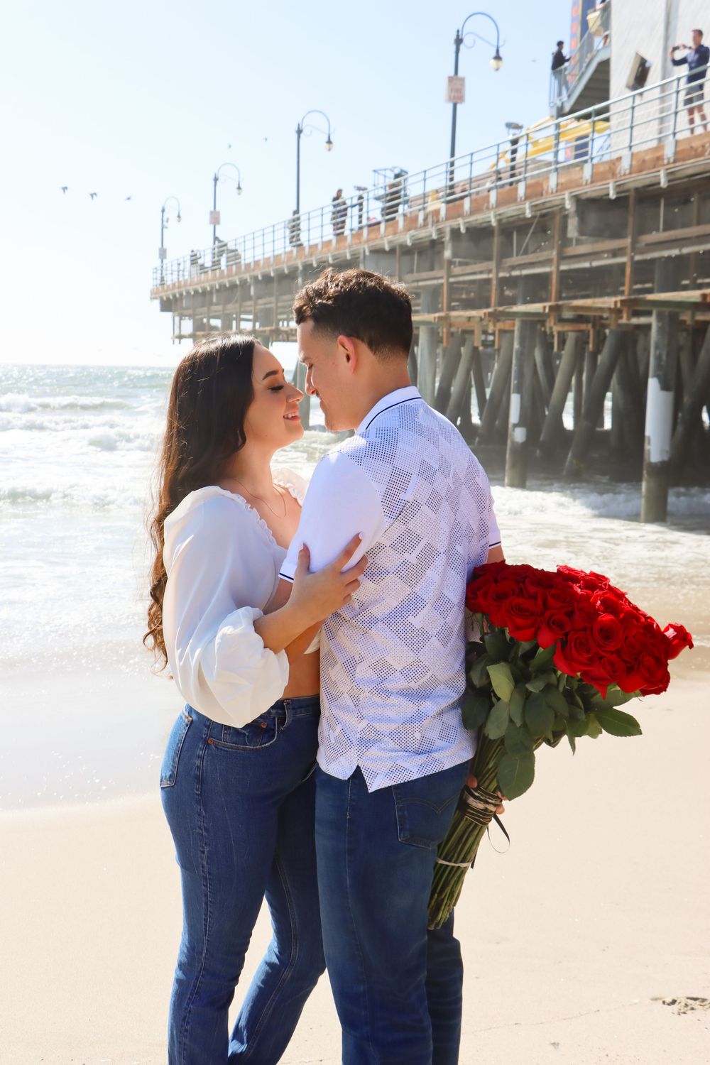 A couple embraces on a beach near a pier while holding red roses.