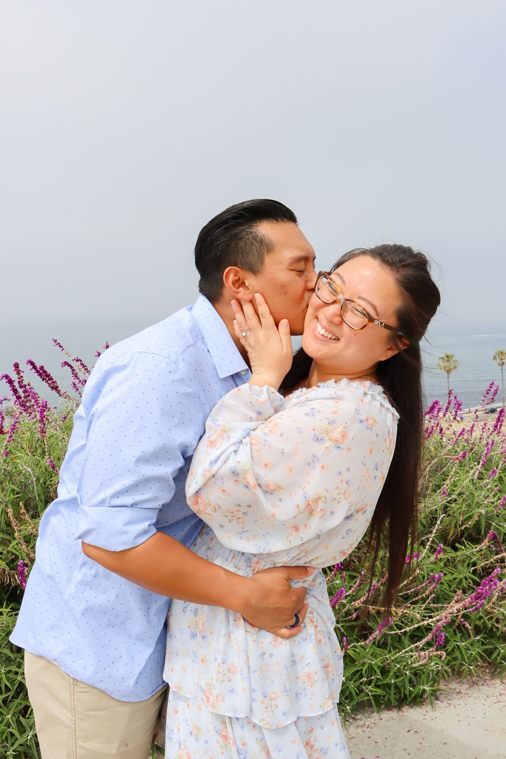 A couple shares affectionate moments among blooming purple flowers by the coast.