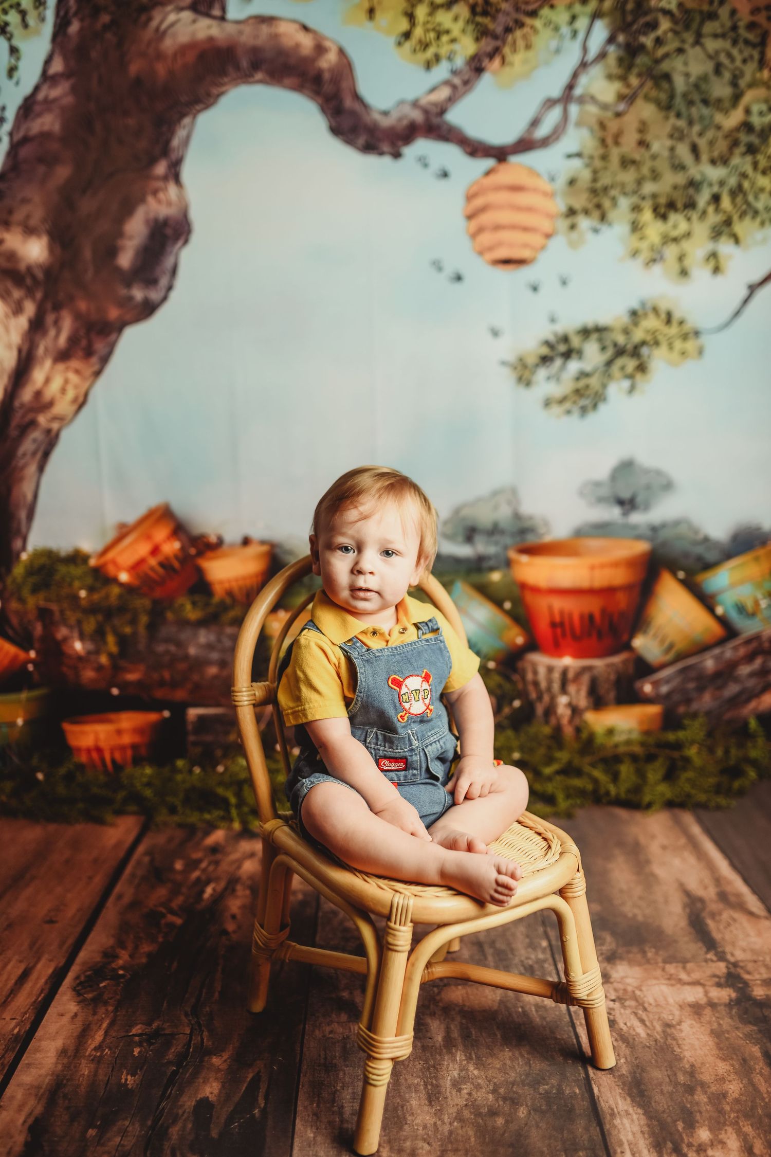 Toddler in denim overalls sits in wicker chair against whimsical woodland backdrop.