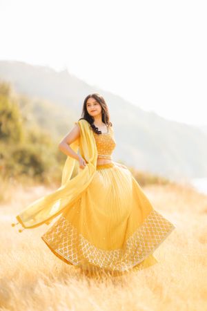 A woman in a flowing yellow traditional dress twirls in a sunlit field with mountains in the background.