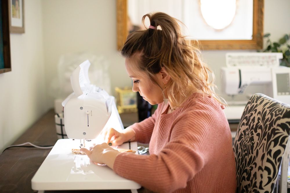 Side view of someone sewing at a white machine in a cozy home studio setting.