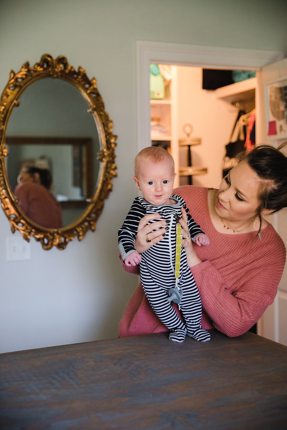 A person in pink holding a baby in striped clothing near an ornate gold mirror.