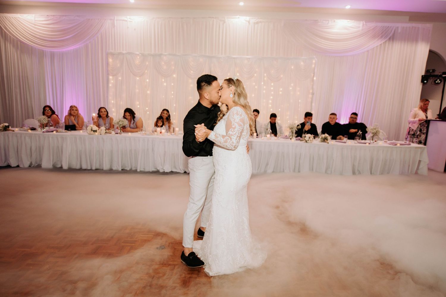 A couple shares their first dance at a wedding reception with guests seated at decorated tables in the background.