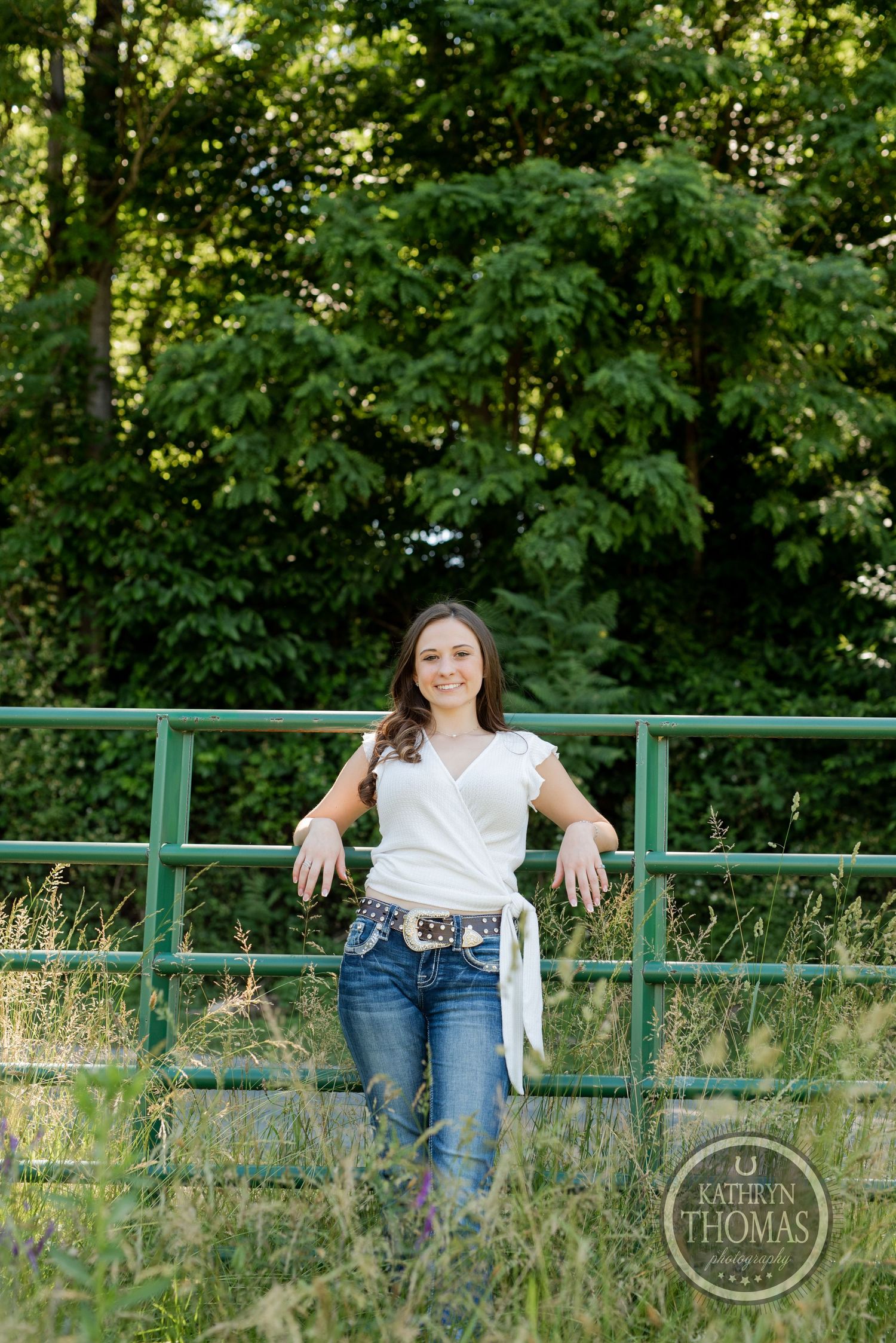 A full-length outdoor portrait against green trees with a decorative green railing.