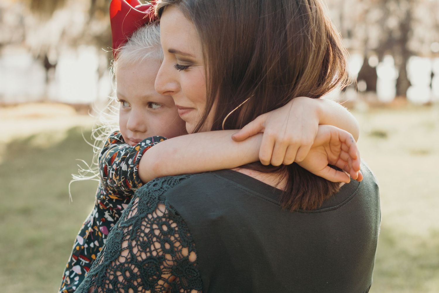 Mom holding and embracing young daughter with both of them looking off. Daughter has arms wrapped around mom's neck.
